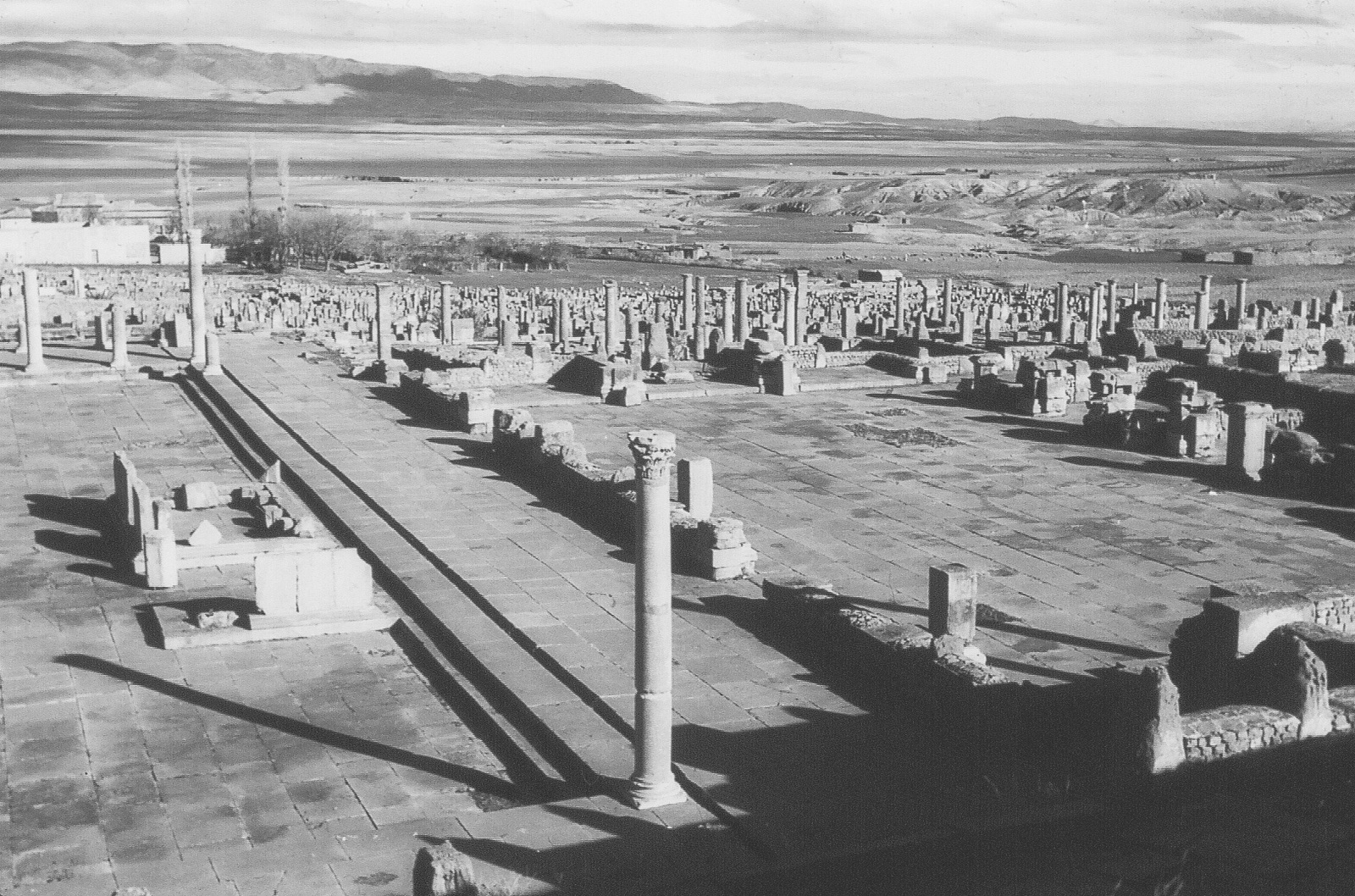 A landscape image depicting the ruins of Timgad, an ancient Roman city. The ruins are surrounded by severly eroded lands, which according to Lowdermilk are now only able to support a few hundred inhabitants living in the huts shown in the background.