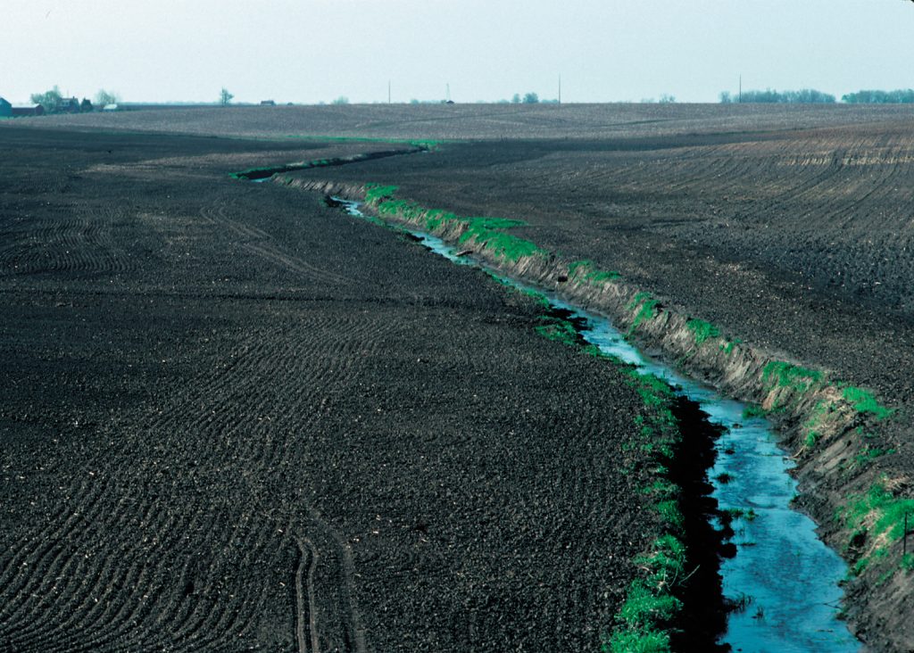 A landscape photograph of a field with bare, black soil that was freshly tilled up to the edge of a stream that disects the landscape through the center of the image. The stream has sparse green plants, but is mostly bare due to recent streambank erosion.