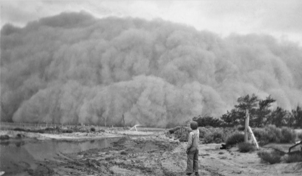 A black and white photograph depicting a young boy standing next to a stream pointing towards an oncoming, ominous-looking dust storm on the horizon. The boy is wearing overalls, a light colored long sleeved shirt, and a dark hat.