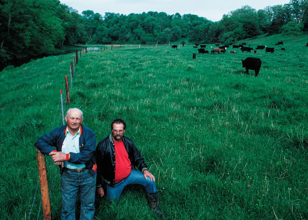 Two men pose next to a barbed wire fence keeping the cattle in the picture out of the nearby stream.
