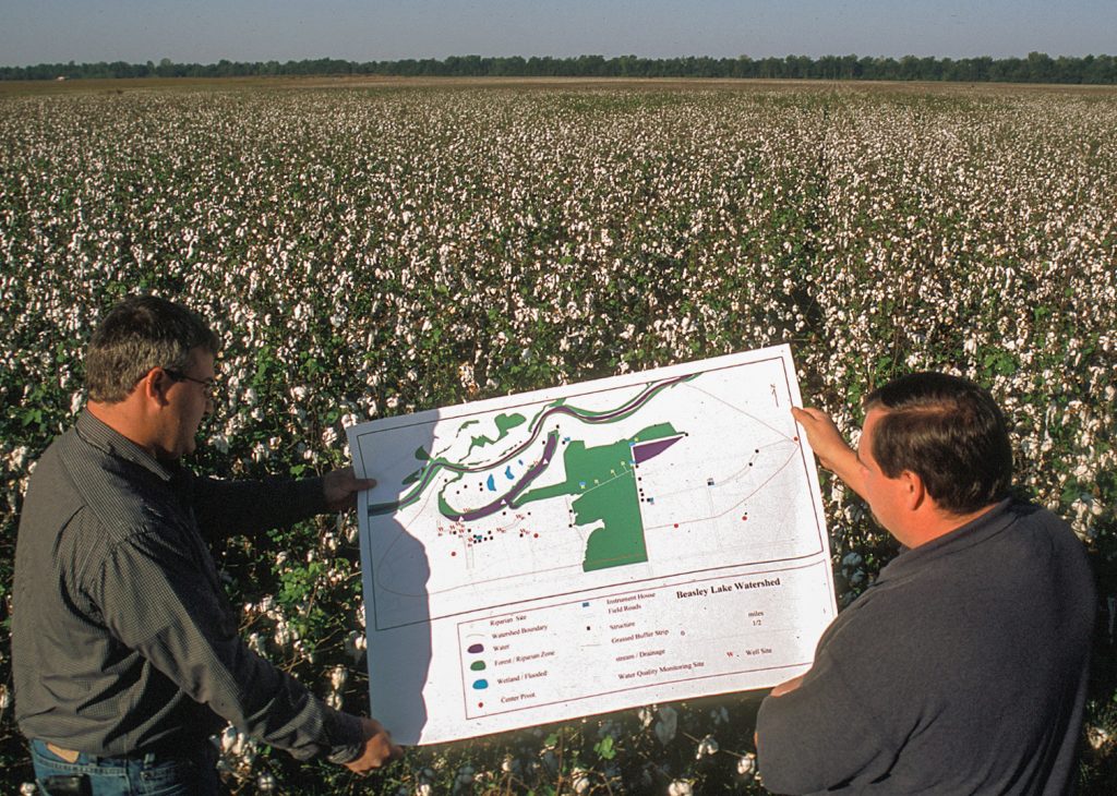 Two men hold up construction plans for a wetland restoration project planned for the cotton field in which they are standing.