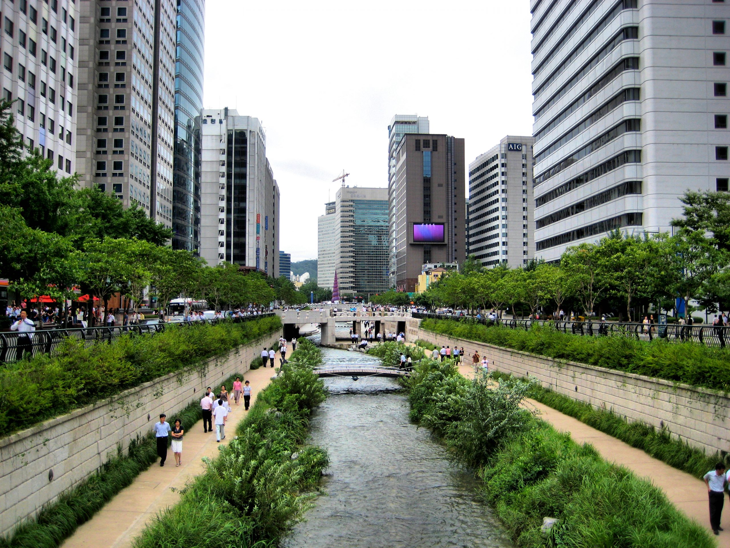 An urban stream lined with pedestrian walkways, retaining walls, city streets, and high rise buildings.