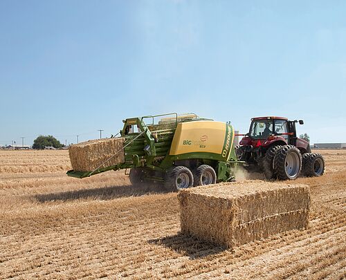 A tractor pulling a square baler that is dispensing a square bale in a hayfield.