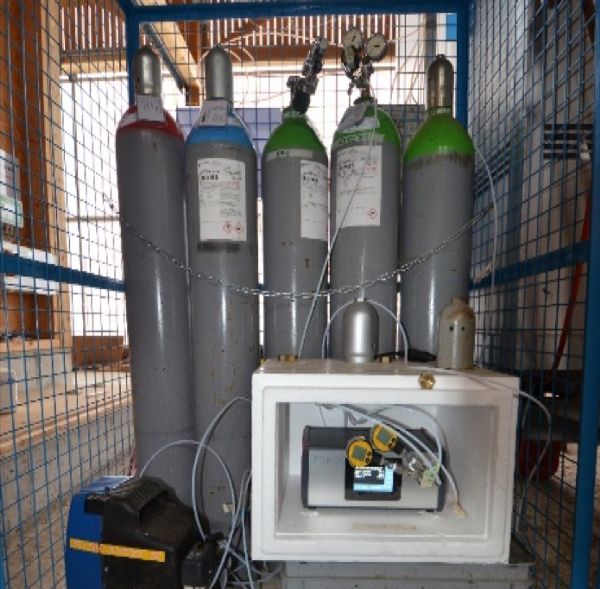 Five gas tanks lined up with a mass-flow controller in front of them and surrounded by a metal cage.