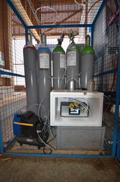 Five gas bottles lined up with a mass-flow controller in front of them and surrounded by a metal cage.