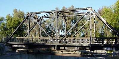 A wooden bridge over a river, consisting of a flat span, several pairs of vertical supports, and a pair of beams parallel to the span, located at the tops of the supports. All of these components are connected by diagonal beams.
