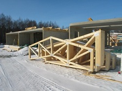 A roof truss made of connected wooden beams which all lie in the same plane, next to a house under construction in a snowy field.