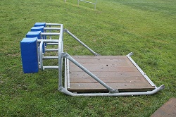 A training sled, consisting of a wooden platform attached on top of a series of metal rods in contact lengthwise with the ground, is on a grassy field.