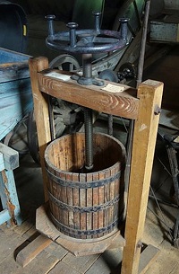 A cider press consisting of a wooden bucket and frame connected by a large metal screw, rotated by turning a large wheel-shaped handle.