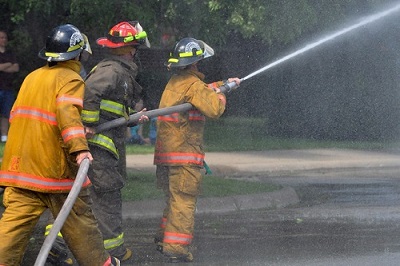 Two firefighters hold the body of a fire hose as it sprays water, while a third firefighter controls the direction of the nozzle of the hose.
