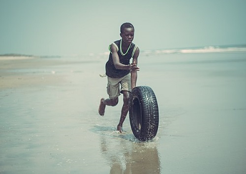 A boy running along a beach, rolling a tire on its side.
