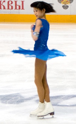 A figure skater in a blue dress doing a spin, with her arms tucked in tight to her body.