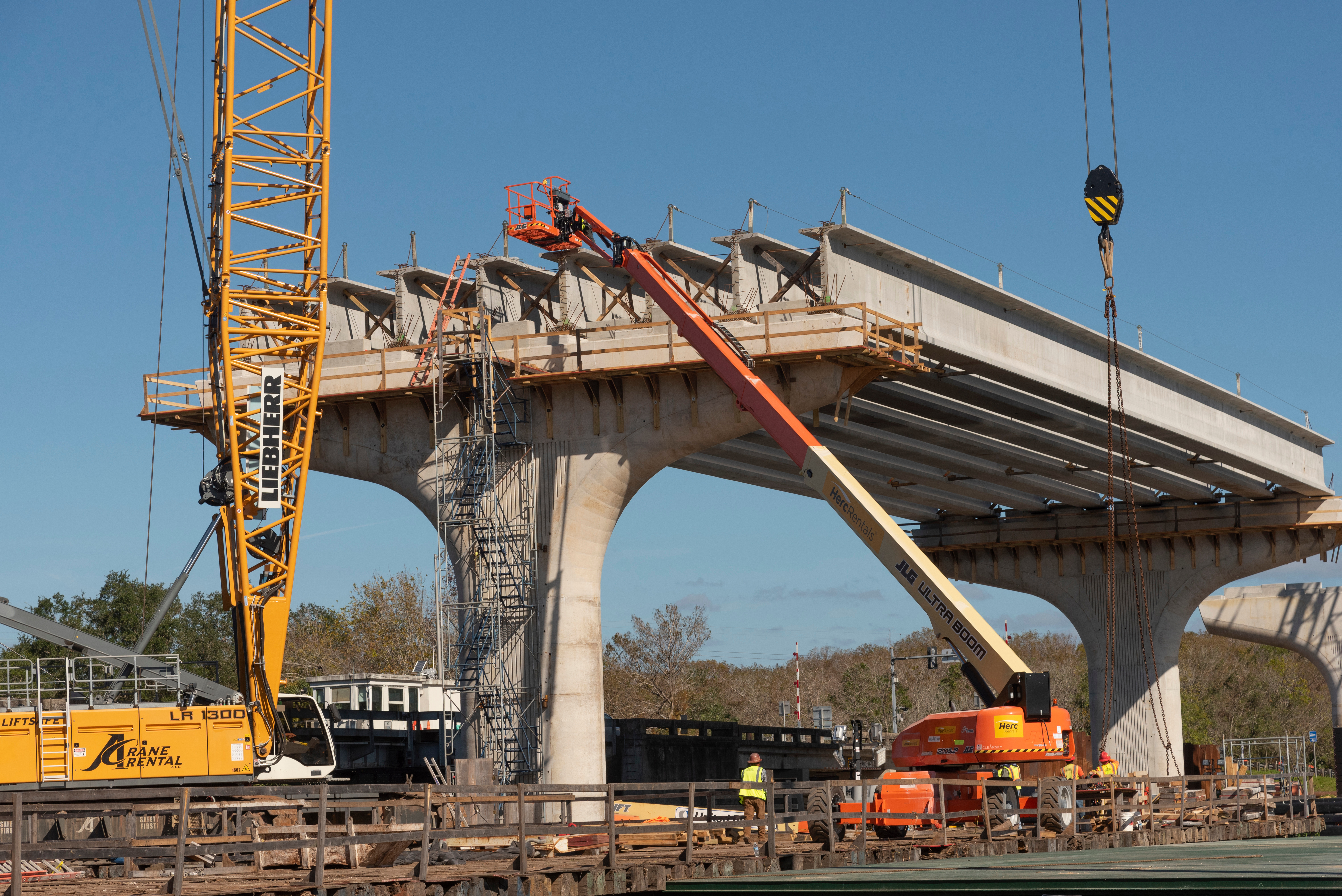 A bridge under construction with cranes and workers using equipment to assemble large concrete beams and supports in a clear outdoor setting.
