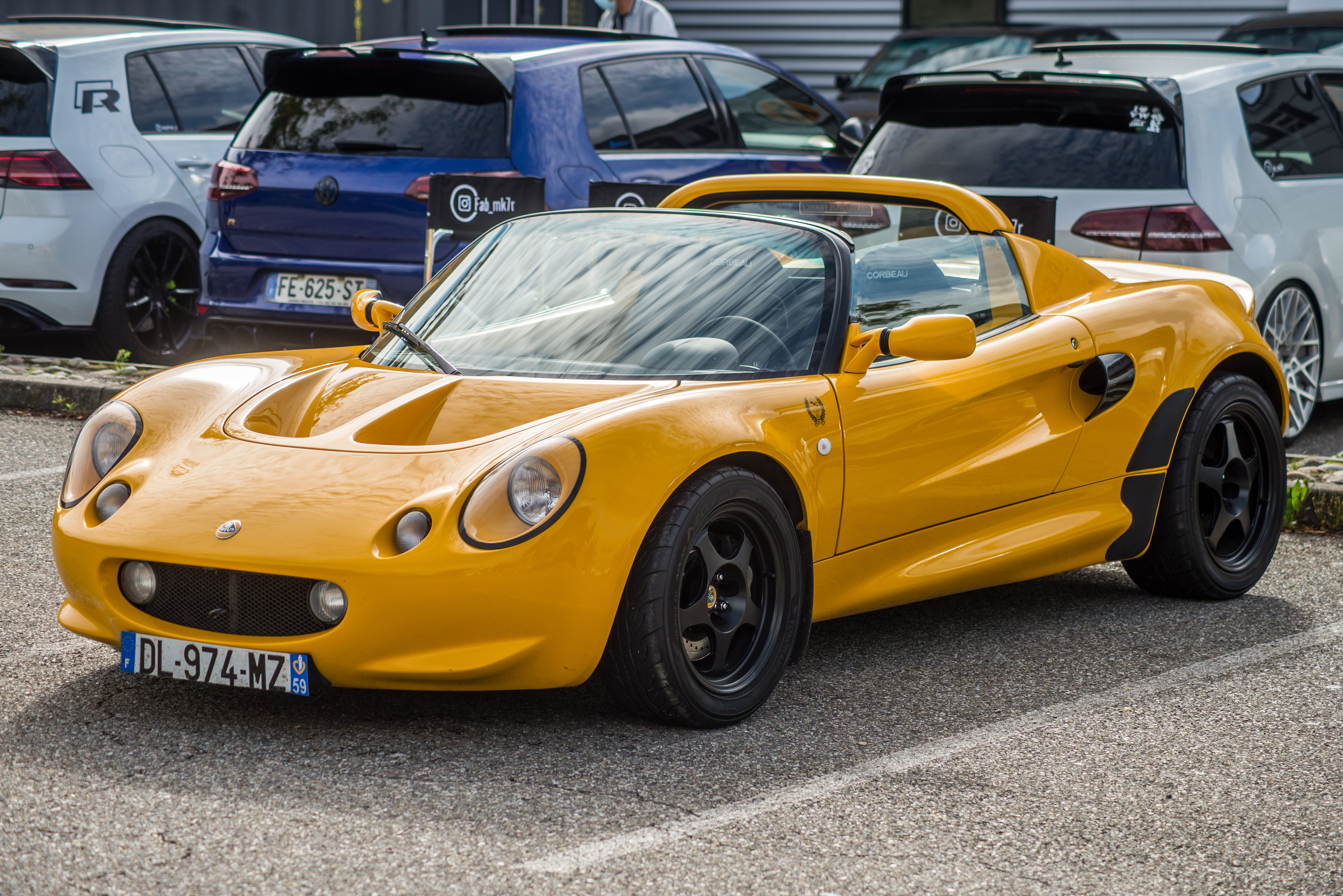 A yellow Lotus Elise sports car parked in a lot with other vehicles in the background, featuring its sleek, aerodynamic design and distinctive round headlights.