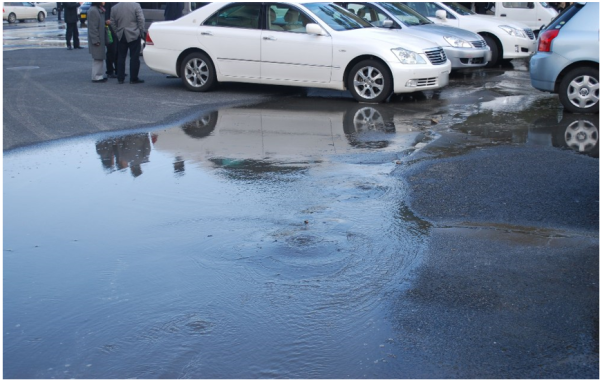 Photograph of the surface of a carpark. There is a large puddle extending from beneath the cars to the foreground of the photo, and the surface of the carpark has settled as result of soil liquefaction.